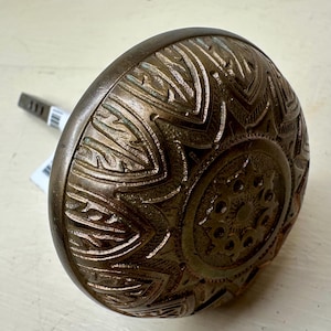 May include: Antique bronze door knob with an ornate, raised floral and geometric pattern. The knob is round with a detailed design and a square spindle. The finish has a weathered appearance.