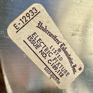 May include: A rectangular sticker with the text "E-12933" in the upper left corner. The sticker reads "Underwriters Laboratories, Inc." and "ELECTRIC FIXTURE" with the issue number "C-189,112". The sticker is on a metallic surface.