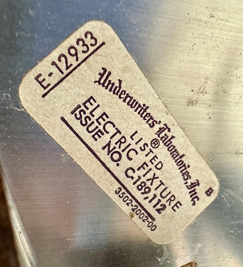 May include: A rectangular sticker with the text "E-12933" in the upper left corner. The sticker reads "Underwriters Laboratories, Inc." and "ELECTRIC FIXTURE" with the issue number "C-189,112". The sticker is on a metallic surface.