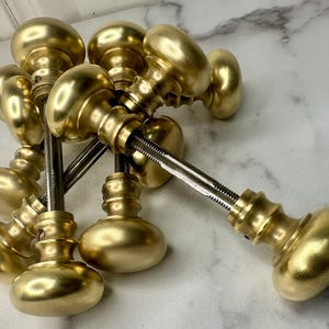 May include: A collection of antique brass door knobs. The knobs have an oval shape and a shiny, golden finish. Each knob is attached to a metal rod with a threaded end. The knobs are arranged in a pile, suggesting a set or collection.