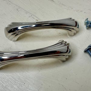 May include: Two polished chrome cabinet handles with a decorative, fluted design. The handles are accompanied by four blue screws, likely for installation. The hardware is set against a light-colored surface.