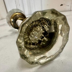 May include: A clear, faceted glass doorknob with a brass base. The knob has an octagonal shape and a detailed interior design. The hardware is designed for a door and is likely vintage.