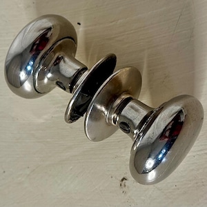 May include: Two polished, oval-shaped door knobs with circular backplates. The knobs are made of a shiny, reflective metal, likely chrome or nickel. The hardware is designed for a door and is likely vintage or antique.