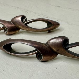May include: Two bronze-colored cabinet pulls with an abstract, flowing design. Each pull features an elongated oval shape with a looped, curved element. The hardware is set against a light-colored, textured surface.