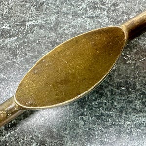 May include: Close-up of a vintage brass spoon with an oval-shaped bowl. The spoon has a tarnished, aged appearance, with a dark, mottled finish. The handle is slender and tapers towards the end.