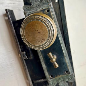 May include: Antique door hardware set featuring a bronze-colored round doorknob with a decorative rim. The set includes a black metal plate with ornate detailing and a matching keyhole cover. The hardware is mounted on a white surface.