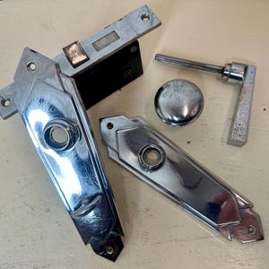 May include: An assortment of vintage door hardware, including a chrome-plated door handle, a door knob, a latch, and two decorative escutcheon plates. The hardware has a shiny, reflective surface and shows signs of age.