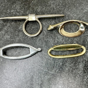 May include: Four metal hardware pieces in silver and gold tones are displayed against a dark, textured surface. The pieces include a ring with a bar, an oval shape, and two different handle designs. These are likely cabinet or furniture hardware.