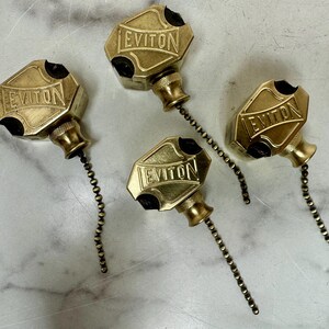 May include: Four vintage brass pull chain light switch pulls. Each pull has a hexagonal shape with the word "LEVITON" embossed on the front. The pulls are connected to a chain of small brass beads.