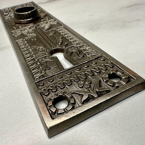 May include: A rectangular, antique-style door plate with intricate embossed patterns. The plate is a muted bronze color and features a keyhole, a circular opening, and decorative floral and geometric designs. The overall aesthetic is vintage.