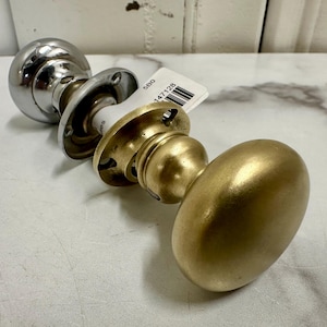 May include: A polished brass door knob with a round, oval-shaped design. The knob is attached to a silver-colored base with a round, flat plate.