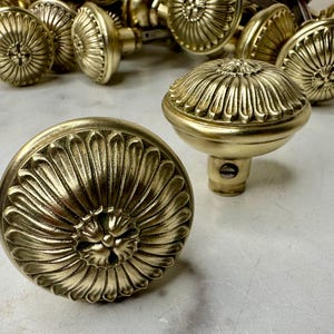 May include: Close-up of several antique brass door knobs. The knobs have a detailed floral design on the round head. The brass has a warm, golden hue, and the knobs are arranged on a light-colored surface.