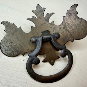 May include: Antique brass drawer pull with a decorative backplate and a curved handle. The backplate has an ornate, leaf-like design. The handle is a dark, aged bronze color. This hardware is suitable for cabinets and furniture.