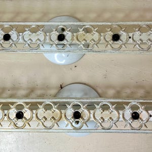May include: Two vintage, off-white metal wall sconces. Each sconce features an ornate, openwork design with a series of quatrefoil shapes and black accents. The sconces have a curved, decorative edge and a circular base.