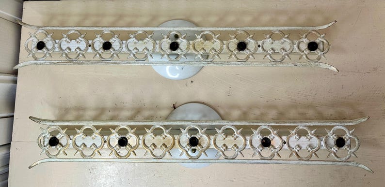 May include: Two vintage, off-white metal wall sconces. Each sconce features an ornate, openwork design with a series of quatrefoil shapes and black accents. The sconces have a curved, decorative edge and a circular base.