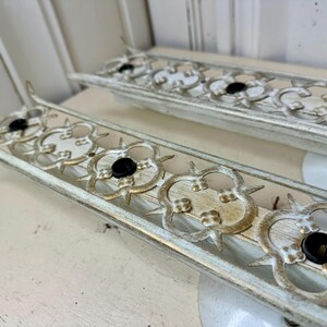 May include: Two vintage, rectangular light fixtures with a distressed white and gold finish. Each fixture features a decorative, openwork design with black bulb sockets. The fixtures are placed on a white surface.