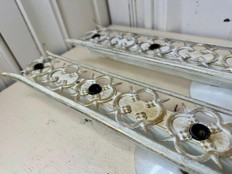 May include: Two vintage, rectangular light fixtures with a distressed white and gold finish. Each fixture features a decorative, openwork design with black bulb sockets. The fixtures are placed on a white surface.