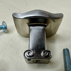May include: A silver-toned cabinet knob with a curved top and a rectangular base. The base features decorative scrollwork. Two screws are visible, one on each side of the knob. This hardware is suitable for furniture and home decor projects.