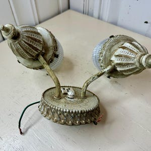May include: An antique wall sconce with two lights. The sconce has a distressed, off-white finish with decorative detailing. Each light has a textured glass shade and a curved arm. The base has a decorative edge and a small switch.