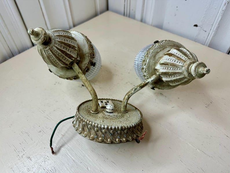 May include: An antique wall sconce with two lights. The sconce has a distressed, off-white finish with decorative detailing. Each light has a textured glass shade and a curved arm. The base has a decorative edge and a small switch.