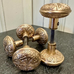 May include: A set of antique brass door knobs with intricate floral and leaf patterns. The knobs are a warm gold color and are arranged on a dark surface, showcasing their detailed design.