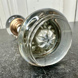 May include: Clear glass doorknob with a silver-colored interior design. The doorknob has a round shape and a decorative floral pattern on the inside. The base is a copper color.