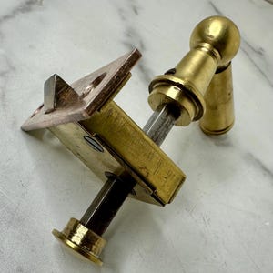 May include: A brass and copper door latch with a rectangular body and a round handle. The latch has a square bolt and a copper strike plate. The hardware is designed for a door and is made of metal.