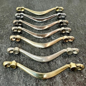 May include: Six curved cabinet pulls in various metallic finishes, including gold, silver, and bronze. The pulls have a classic, arched design with cylindrical end pieces for mounting. They are arranged on a dark, textured surface.