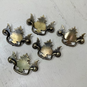 May include: Five antique brass drawer pulls with a leaf design. The pulls have a curved handle and decorative accents. The brass has a warm, aged patina, adding to their vintage charm. These pulls are ideal for adding a touch of elegance to furniture.
