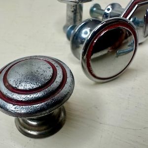 May include: A collection of vintage cabinet knobs. The knobs are primarily silver with red accents. One knob has a circular design with concentric red rings. The knobs have a distressed, aged appearance, suggesting they are antique or vintage.