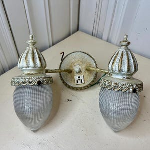 May include: Vintage double-light wall sconce with a white and gold finish. The sconce features two clear, textured glass globes and ornate detailing. The fixture includes a circular backplate and a built-in electrical outlet.