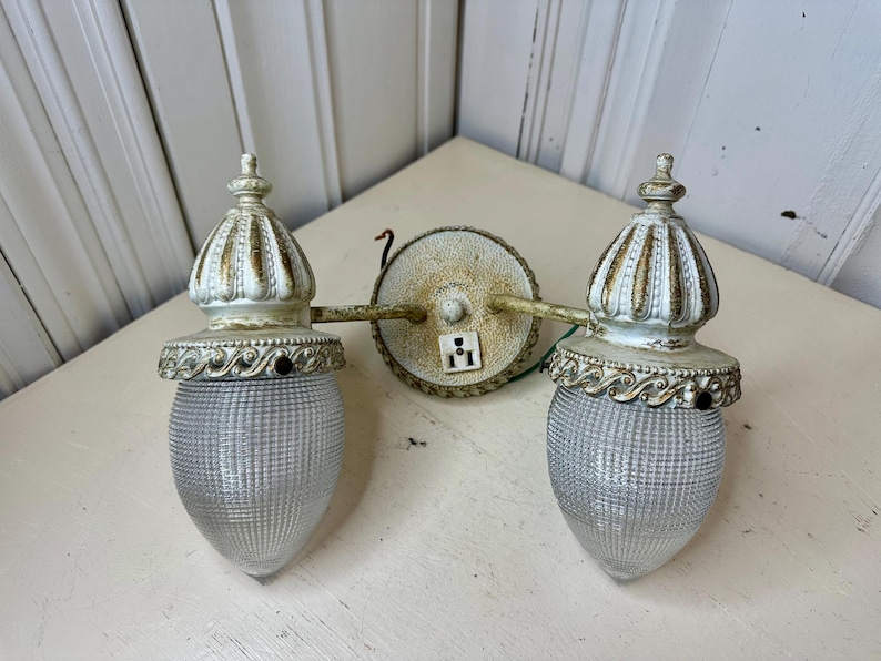 May include: Vintage double-light wall sconce with a white and gold finish. The sconce features two clear, textured glass globes and ornate detailing. The fixture includes a circular backplate and a built-in electrical outlet.
