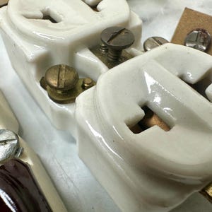 May include: Close-up of vintage electrical components. The image features several white ceramic insulators with brass and steel hardware, including screws and metal contacts. The components have a glossy finish and are arranged on a light-colored surface.