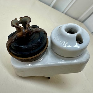 Puede incluir: Componente eléctrico antiguo con base de cerámica blanca. Tiene un componente negro y acanalado con una abrazadera de metal oxidado y una pieza de cerámica blanca con tres agujeros. Objeto vintage.