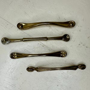 May include: Four vintage brass drawer pulls in varying designs. The pulls are arranged horizontally against a white background. Each pull has a different shape, with rounded or decorative ends and a central bar for gripping. Ideal for furniture restoration.
