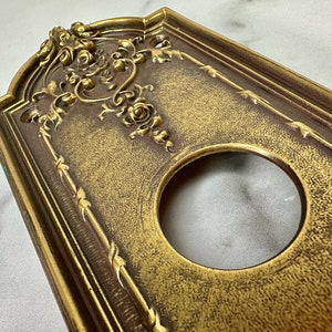 May include: A gold-toned metal door plate with an ornate floral design and a circular cutout. The plate has a textured surface and a slightly aged appearance.
