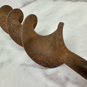 May include: A rusty, antique auger bit with a spiral design. The tool is made of metal and has a long, cylindrical shaft with a curved cutting edge. The auger bit is designed for boring holes in wood.