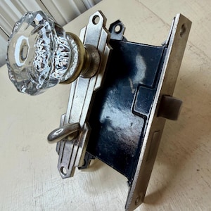 May include: An antique door lock set featuring a clear, faceted glass doorknob. The metal components have a silver finish with a black backing. The lock set has a vintage, Art Deco style.