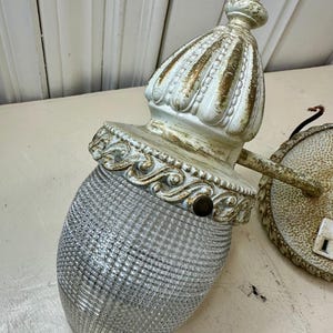 May include: Antique white and gold wall sconce with a textured glass shade. The sconce has a decorative, ornate design with a detailed top and base. The glass shade is a clear, textured, oval shape.