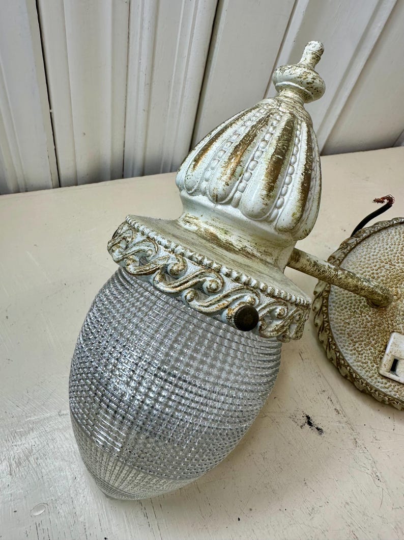May include: Antique white and gold wall sconce with a textured glass shade. The sconce has a decorative, ornate design with a detailed top and base. The glass shade is a clear, textured, oval shape.