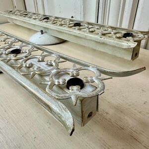 May include: Two vintage, distressed, off-white metal light fixtures with ornate floral designs and black bulb sockets. The fixtures have curved ends and are mounted on white, saucer-shaped bases.