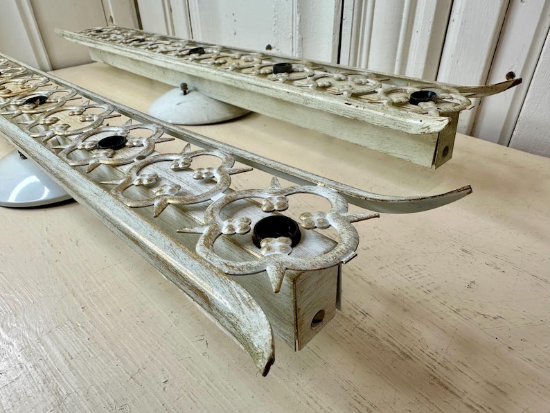 May include: Two vintage, distressed, off-white metal light fixtures with ornate floral designs and black bulb sockets. The fixtures have curved ends and are mounted on white, saucer-shaped bases.