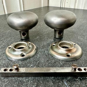 May include: Two antique metal door knobs with matching backplates and a connecting bar. The knobs are a rounded shape with a brushed metal finish. The backplates are circular with screw holes.
