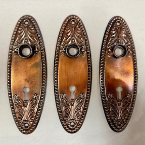 May include: Three antique copper-toned door plates. Each oval plate features a keyhole, a central circular opening, and ornate floral and beaded detailing. The plates have a vintage aesthetic, with a slightly aged patina.