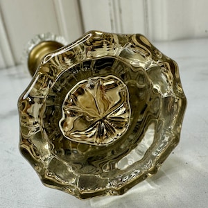 May include: A close-up of a vintage, octagonal-shaped glass doorknob. The knob is a golden color and has a detailed, textured surface. The base is brass-colored. The doorknob is likely for a cabinet or door.