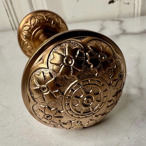 May include: Close-up of an antique bronze door knob. The knob features an ornate floral design with detailed patterns. The bronze finish gives it a vintage look. The door knob is a decorative hardware element.