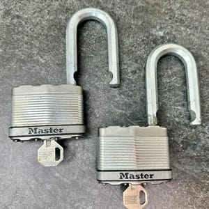 May include: Two silver Master padlocks with open shackles and keys inserted. The locks have a ridged design and the word "Master" is printed on the front. The keys are silver and have a unique keyhole design.
