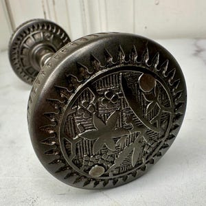 May include: Antique, dark metal door knob with an intricate floral and leaf design. The knob features a circular face with detailed engravings, surrounded by a decorative border. The stem of the knob is also visible in the background.