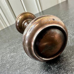 May include: A vintage, antique door knob with a tarnished, aged appearance. The knob is made of metal, with a round, bulbous shape and a stem connecting to a smaller, round base. The metal has a mix of bronze and silver tones, showing signs of wear.