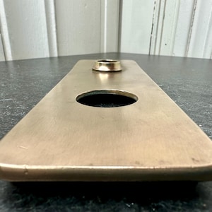 May include: A rectangular bronze-colored metal object with rounded corners. It features a large circular hole and a smaller raised circular element. The surface has a brushed texture, and the object appears to be a decorative or functional piece.